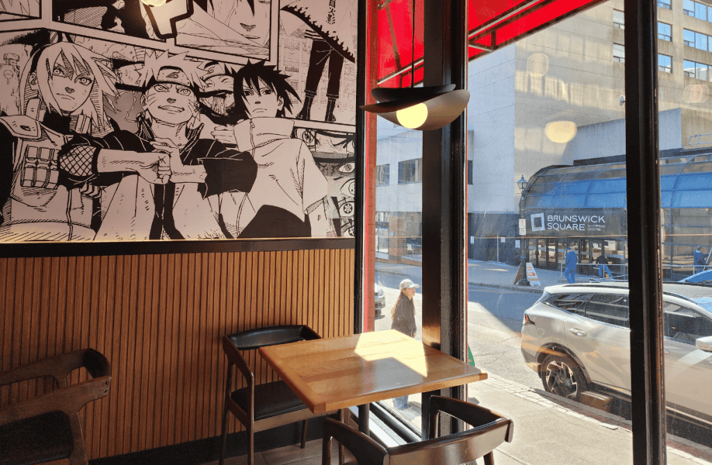 A cozy window-side table at Umami Kid in Uptown Saint John, with manga-style artwork on the wall and views of Brunswick Square across Germain Street.