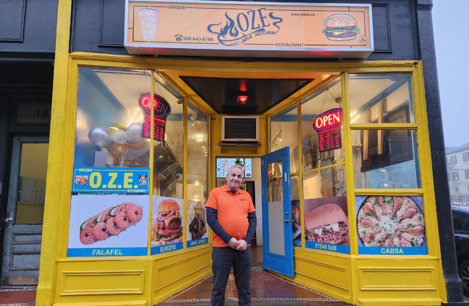 Owner Ahmad Alhaj standing in front of Unique OZE Restaurant at 149 Union Street in Uptown Saint John, with bright yellow storefront, food posters, and an open door inviting customers inside.