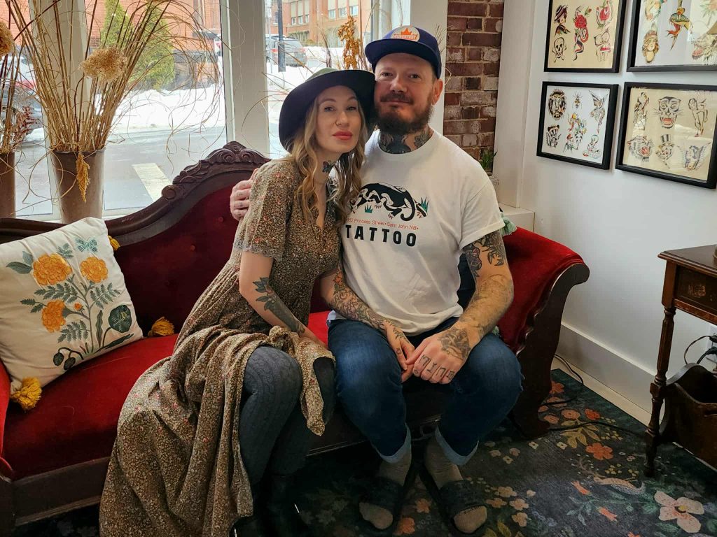 Owners of 162 Princess Street Tattoo, Lenny Johnson and George Keclík, seated together on a vintage red sofa inside their Uptown Saint John studio, surrounded by framed tattoo art.
