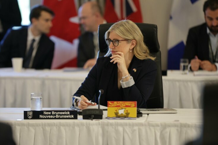 Premier Susan Holt sitting behind a table with a microphone and the provincial flag of New Brunswick